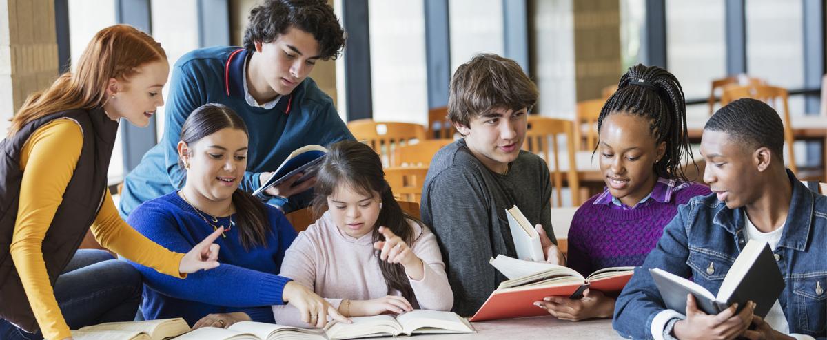 Image of a diverse group of middle school and high school studets sitting together at a table in a library, studying from textbooks and interracting with one another..